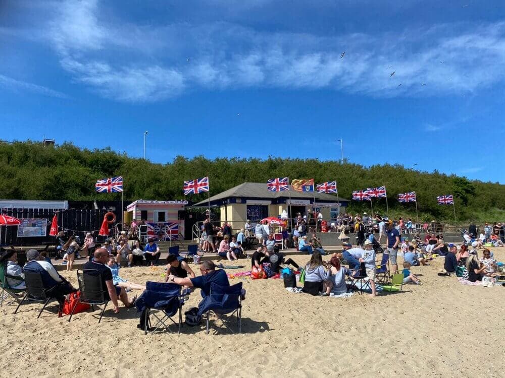 People relaxing on a sunny beach with Union Jack flags displayed and a small building in the background. - Home Instead