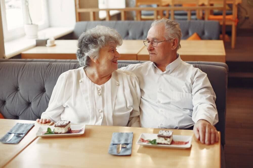 An elderly couple sits closely at a table in a cafe, smiling at each other with dessert plates in front of them. - Home Instead