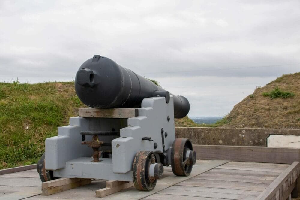 A historic black cannon mounted on a wooden platform with grassy mounds in the background under a cloudy sky. - Home Instead