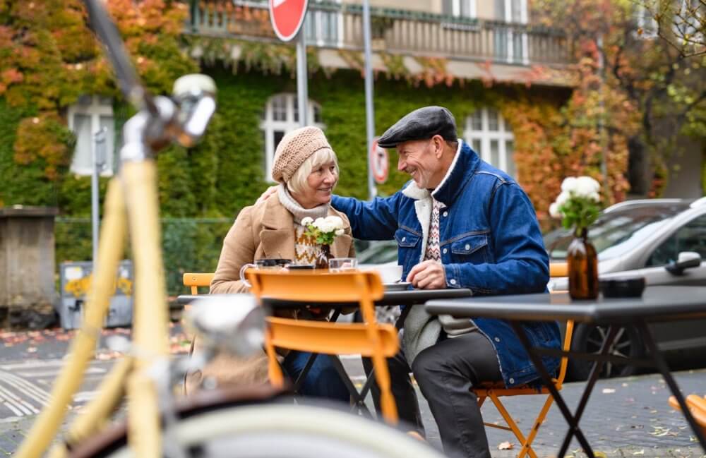 Elderly couple smiling and enjoying coffee at an outdoor café in a cosy, autumn setting. - Home Instead Southampton