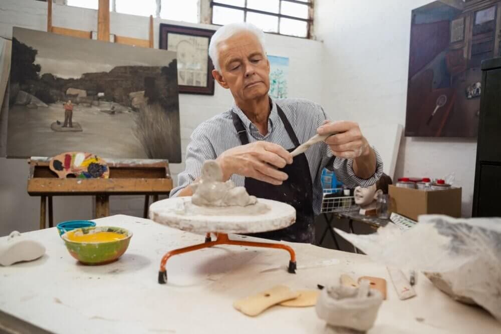 Elderly man sculpting clay in an art studio, with paintings and art supplies in the background. - Home Instead Bournemouth & Christchurch