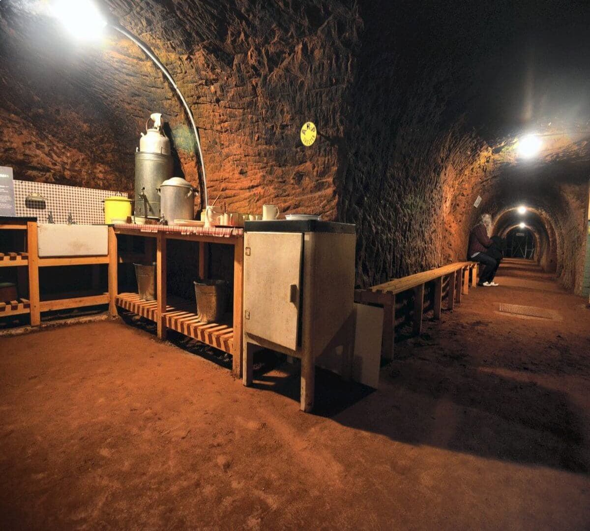 A tunnel with a kitchen area on the left, featuring shelves, a cabinet, and a table; a person sits in the distance. - Home Instead
