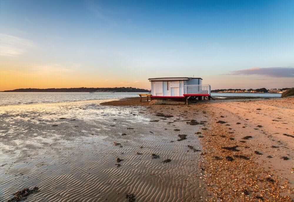 Small beach hut on a sandy shore at sunset, with calm sea water and a clear sky in the background. - Home Instead Poole
