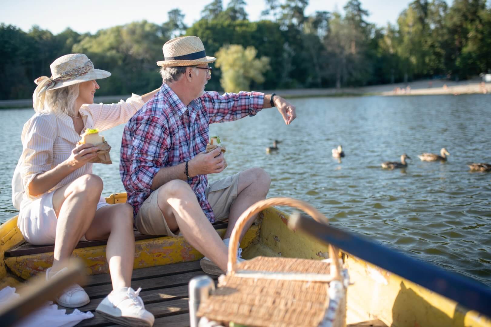 An elderly couple in hats sits on a boat, enjoying drinks and pointing at ducks swimming in a lake. - Home Instead