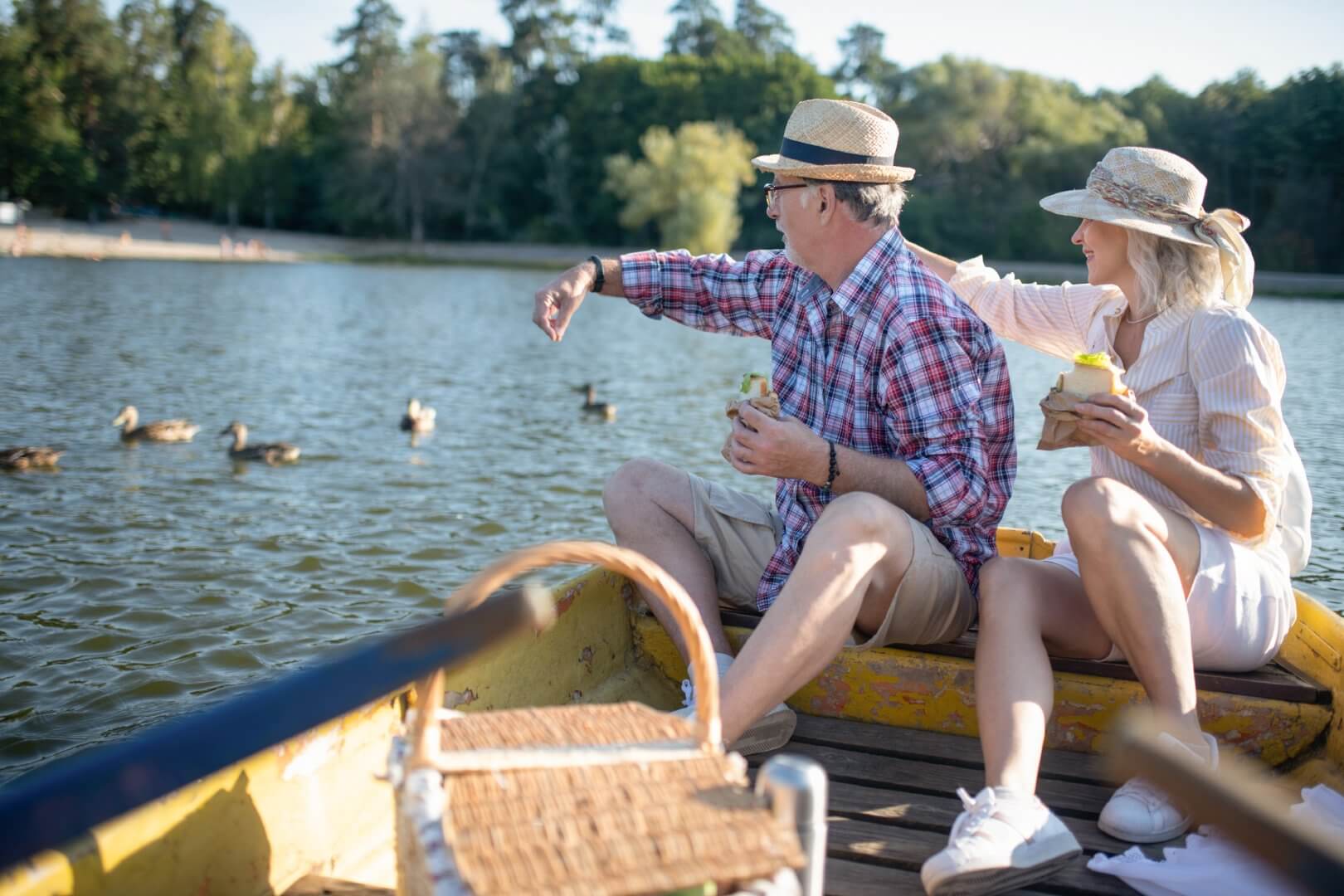 A couple in hats enjoy feeding ducks while sitting on a boat on a sunny day in a lake surrounded by trees. - Home Instead