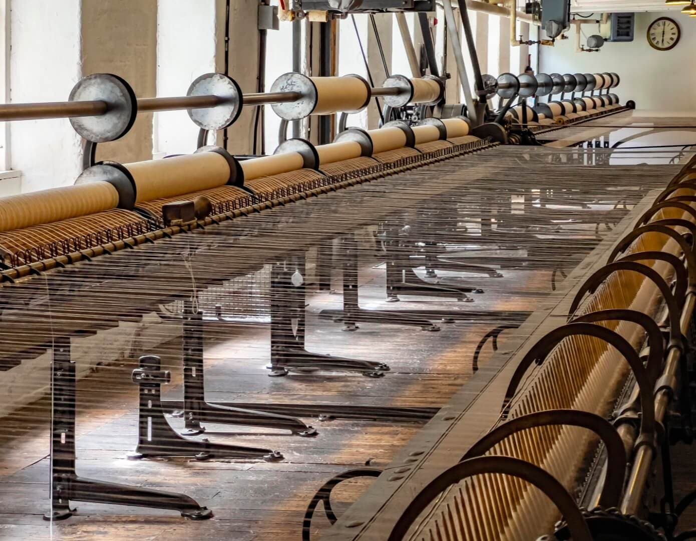 A long row of spinning machines in a historic textile mill, with threads stretching across and a clock on the wall in the background. - Home Instead