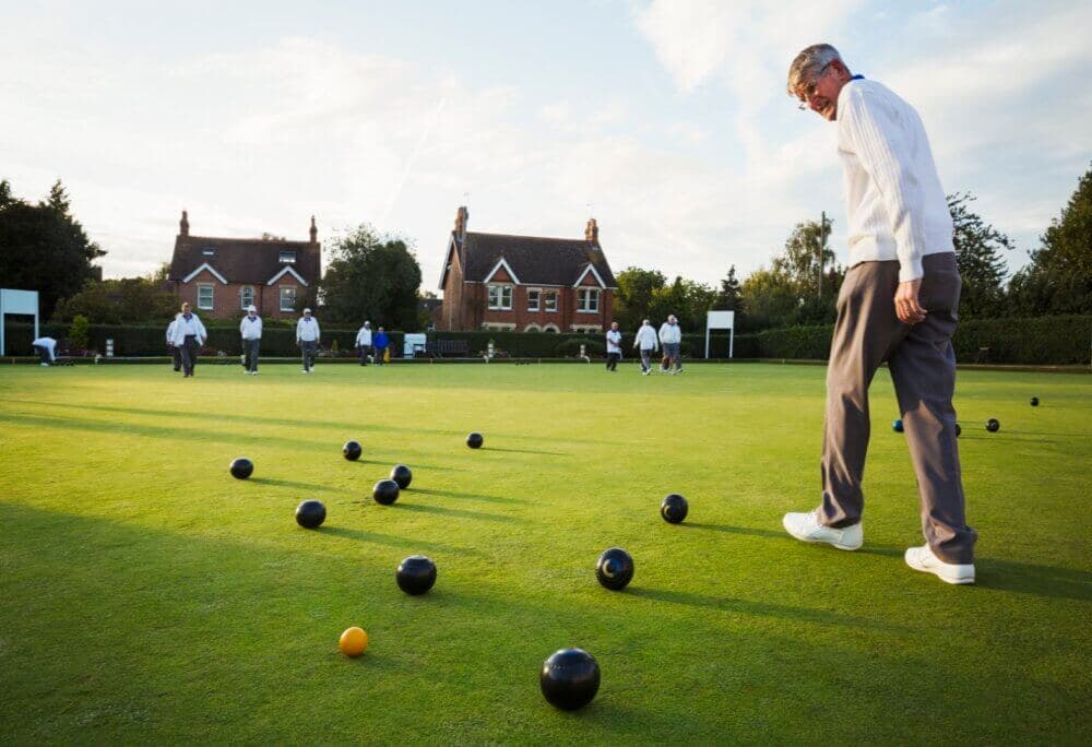A man playing bowls on a green field, with several people and houses in the background. - Home Instead Poole