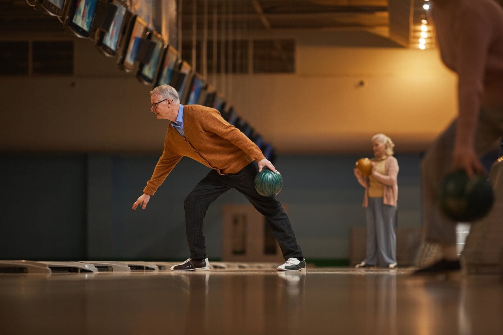 Older man bowling in a dimly lit bowling alley, with a woman in the background holding a bowling ball. - Home Instead Poole