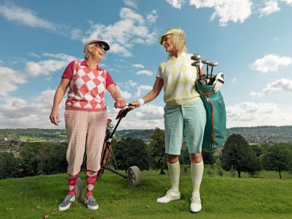 Two elderly women in colorful golf attire enjoying a conversation on a golf course with a scenic background. - Home Instead