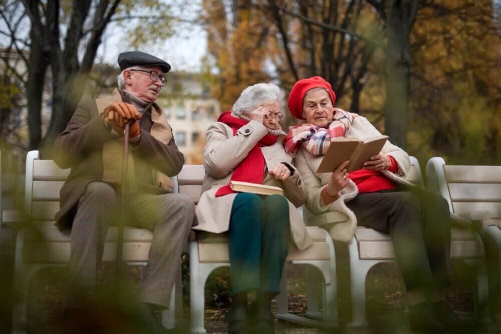 Three elderly friends sit on park benches with books; two women, both in red hats, share a book. The man holds a cane. - Home Instead Bournemouth & Christchurch