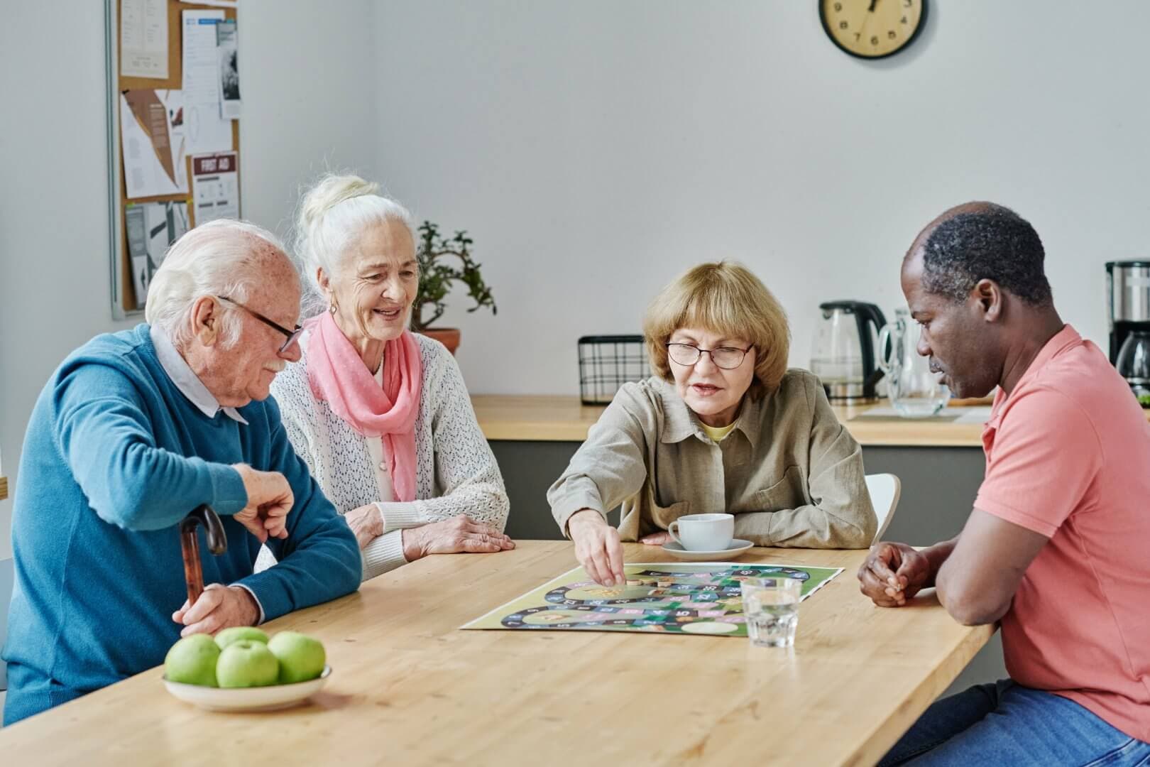 Four elderly people playing a board game together at a wooden table, with apples and a clock in the background. - Home Instead Bournemouth & Christchurch