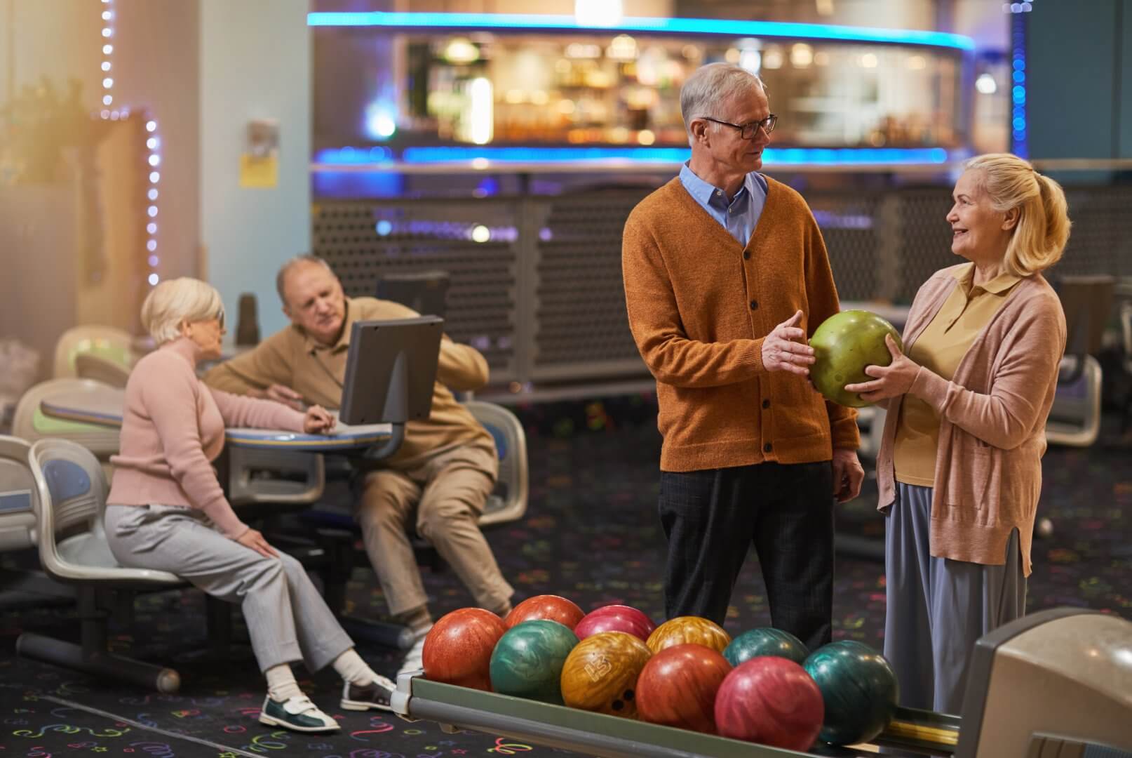 Elderly people bowling, with a couple holding a ball while others sit and chat in a bowling alley. - Home Instead