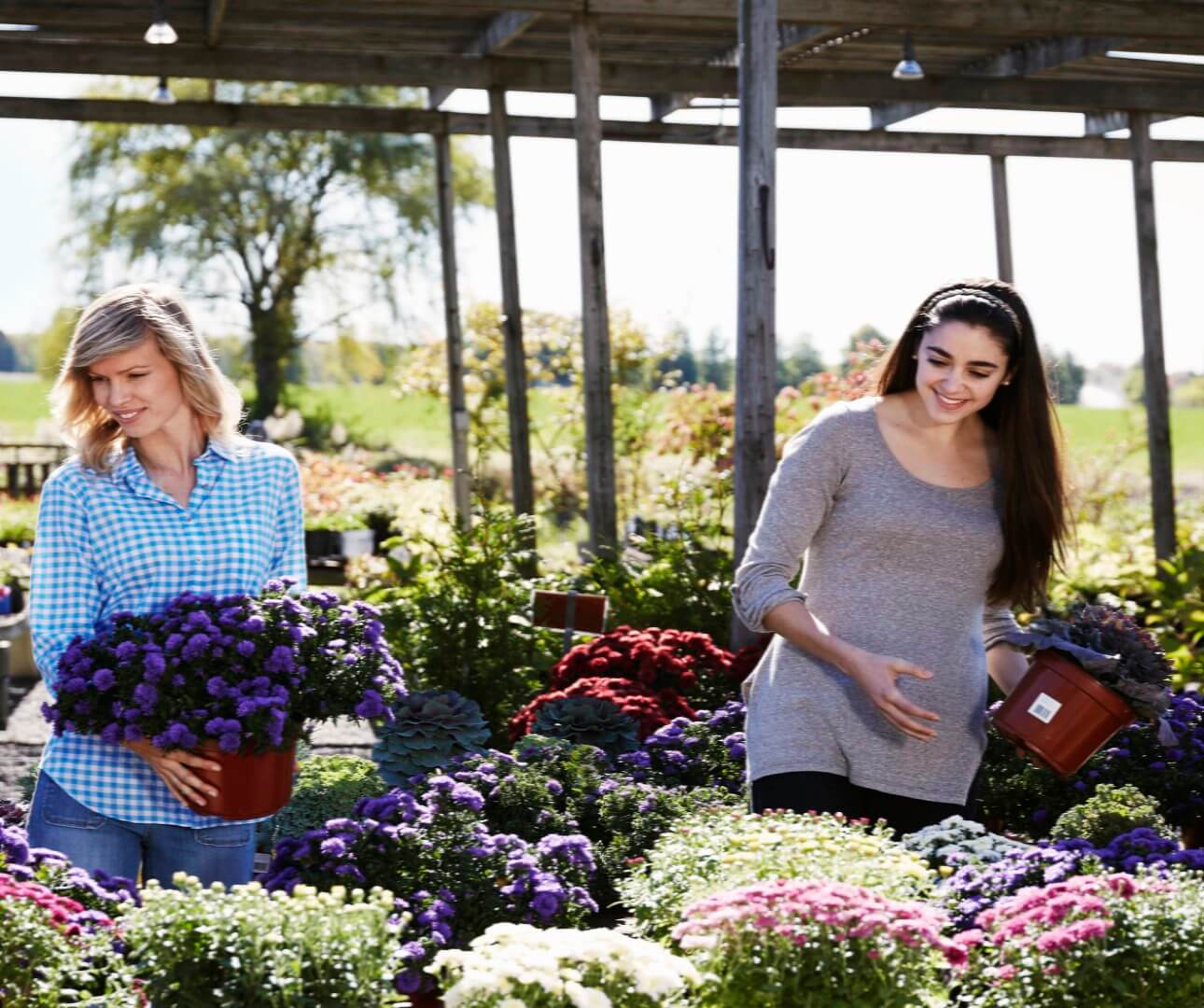 Two women smiling and holding potted flowers at an outdoor nursery, surrounded by colorful plants and flowers. - Home Instead