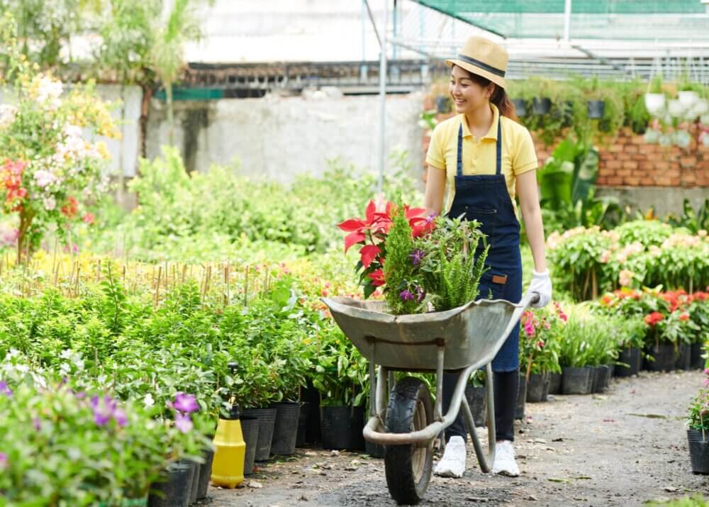 A woman in a hat and apron pushes a wheelbarrow filled with flowers through a garden centre. - Home Instead Southampton