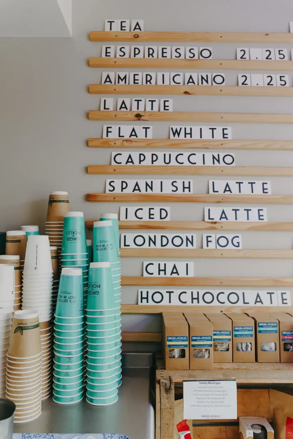 Coffee shop menu with drinks and prices. Stacked paper cups and lids are visible below the menu on the counter. - Home Instead
