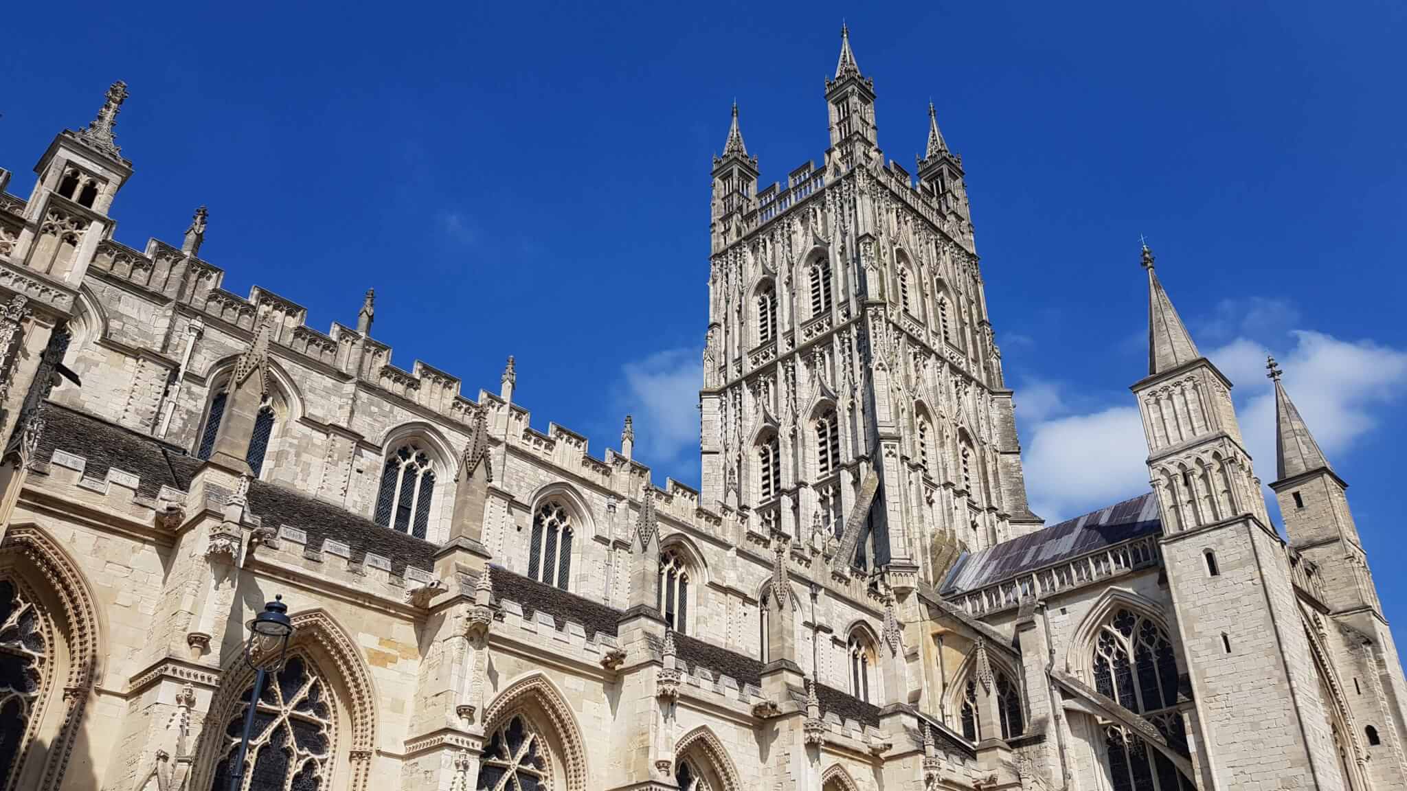 A view of the detailed gothic stonework and tall spires of Gloucester Cathedral against a blue sky. - Home Instead