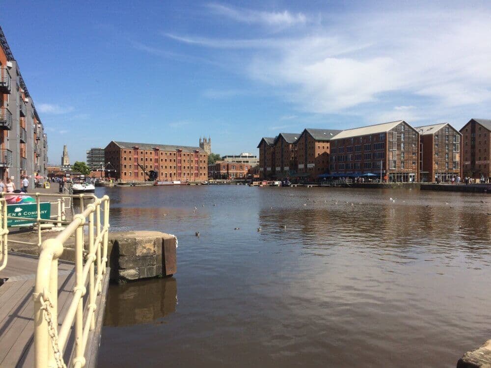 Waterfront scene of Gloucester Docks buildings, boats, and clear blue sky, reflecting in the calm water. - Home Instead