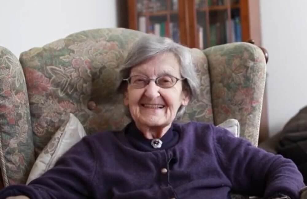 An elderly woman with gray hair and glasses, smiling while seated in an armchair. - Home Instead