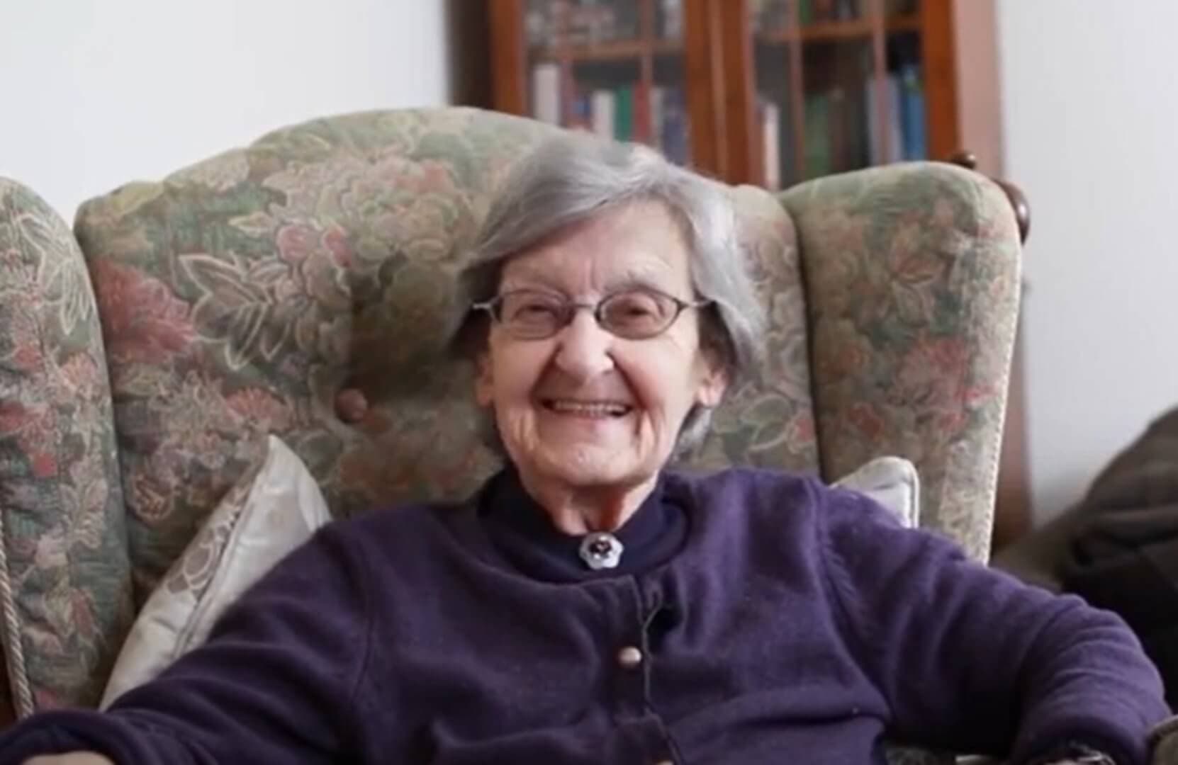 An elderly woman with gray hair and glasses, smiling while seated in an armchair. - Home Instead