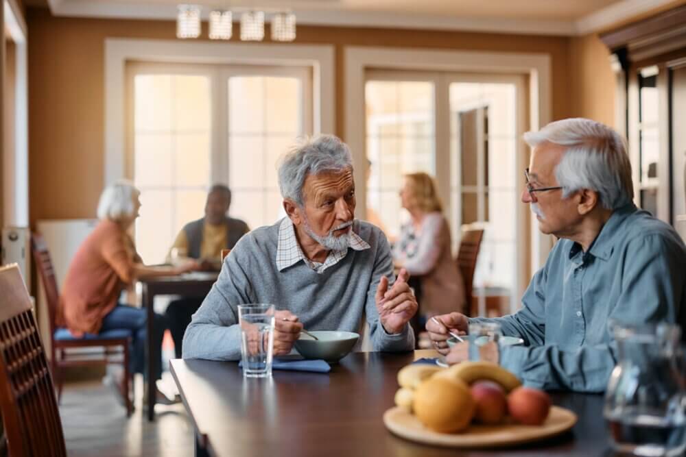 Two elderly men converse over a meal at a dining table with other people sitting at tables in the background. - Home Instead