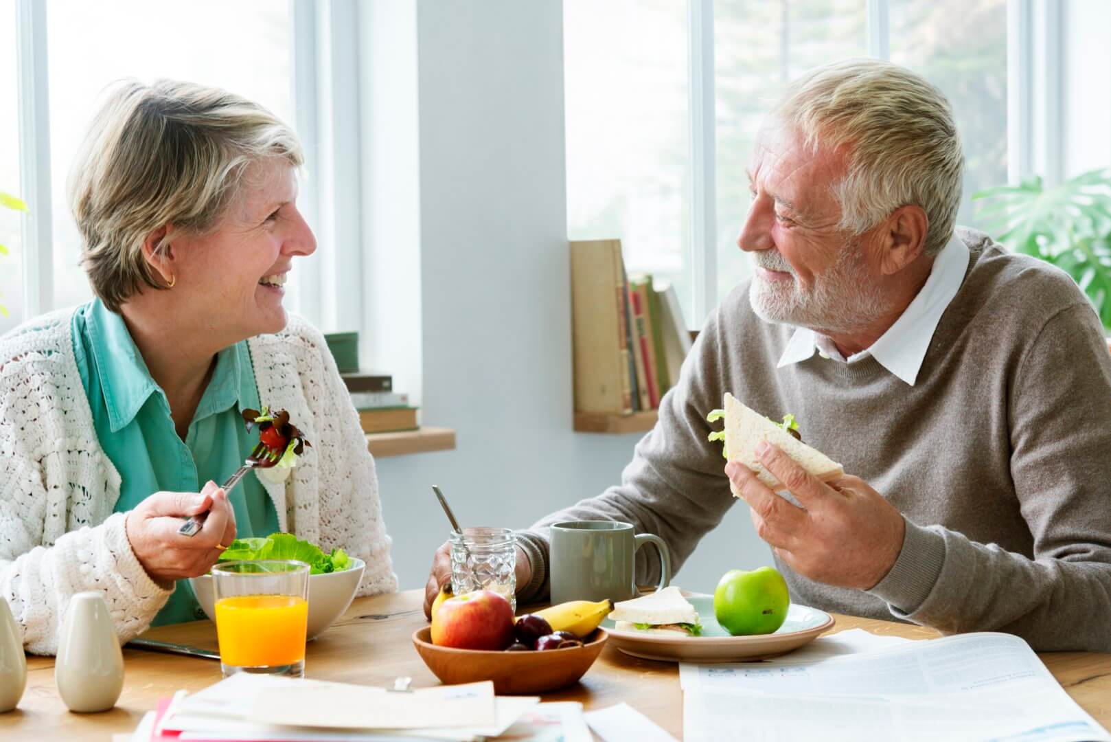 An elderly couple is smiling and eating a meal together at a table with various foods, including salad, fruit, and drinks. - Home Instead