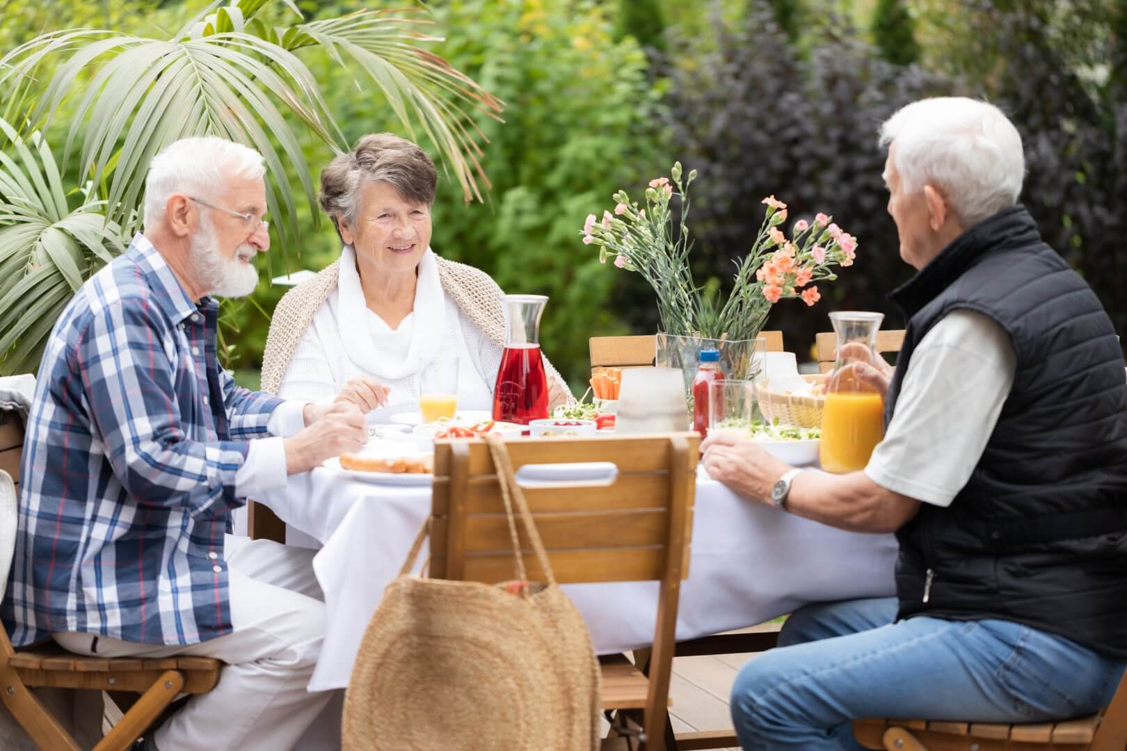 Three elderly friends enjoying a meal at an outdoor table, surrounded by greenery and flowers. - Home Instead Bournemouth & Christchurch