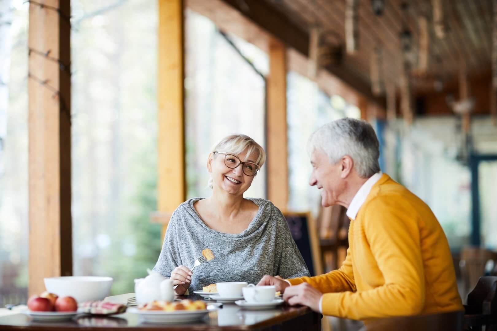 Two older adults sitting and smiling at each other while enjoying a meal at a cosy, sunlit café. - Home Instead Poole