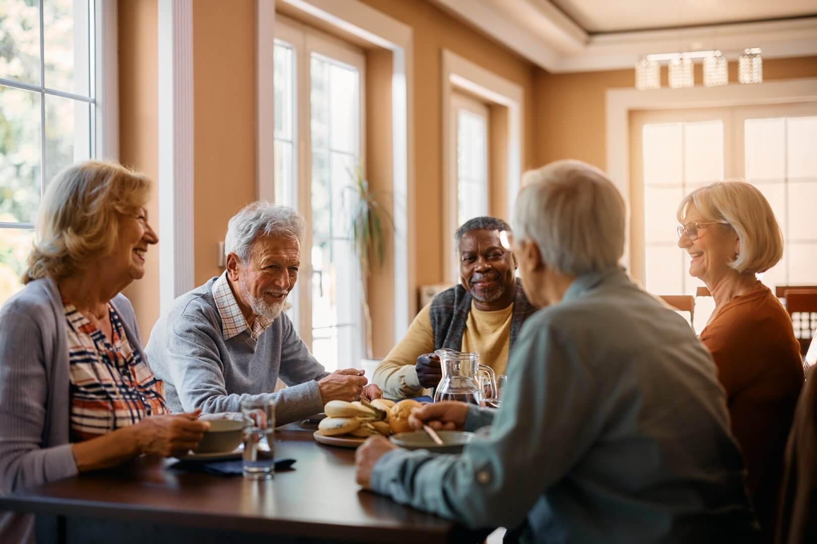 A group of elderly people sit around a table, smiling and chatting in a well-lit room with large windows. - Home Instead