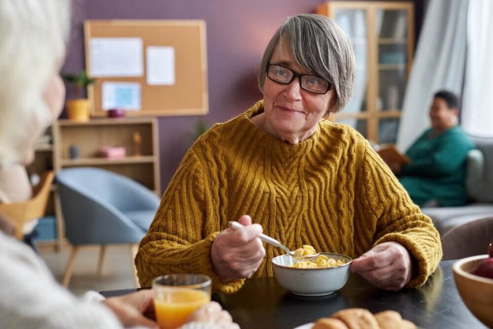 Elderly woman in a yellow sweater eating cereal at a table with other people in a cozy, communal setting. - Home Instead