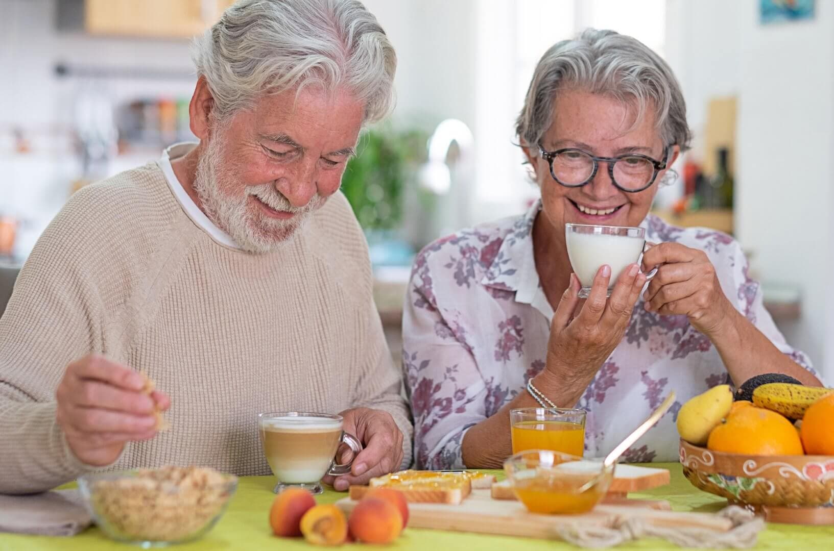 An elderly couple enjoying breakfast together at a table filled with fruit, cereal, and drinks. - Home Instead