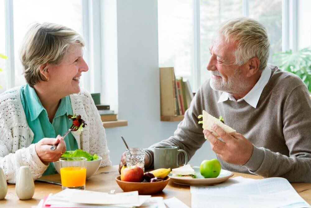Elderly couple sitting at a table, smiling at each other while eating a meal with salad, fruit, and juice. - Home Instead