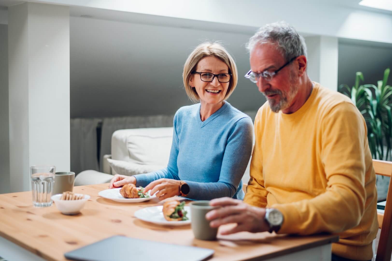 A smiling older woman and man enjoy a meal together at a wooden table in a bright room. - Home Instead