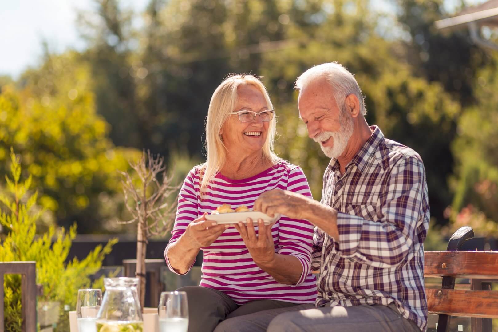 Smiling elderly couple enjoying a meal together outdoors on a sunny day, surrounded by greenery. - Home Instead