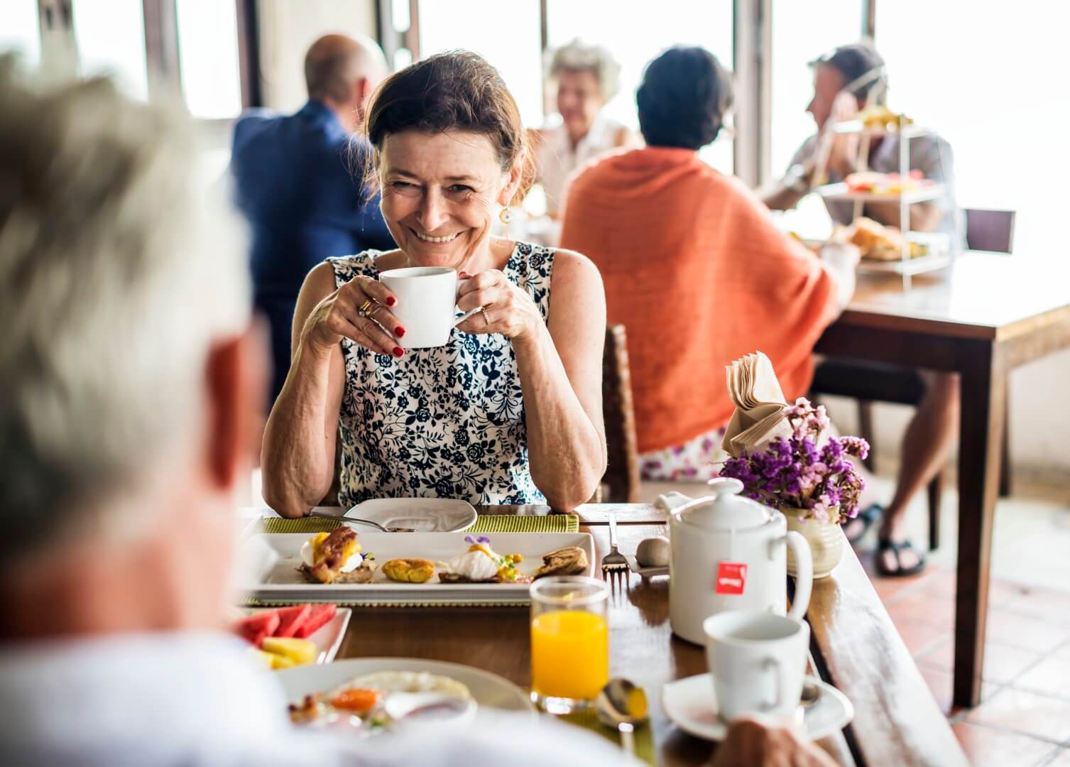 A woman smiles while drinking coffee at a table with a man. Other people are seated at tables in the background. - Home Instead