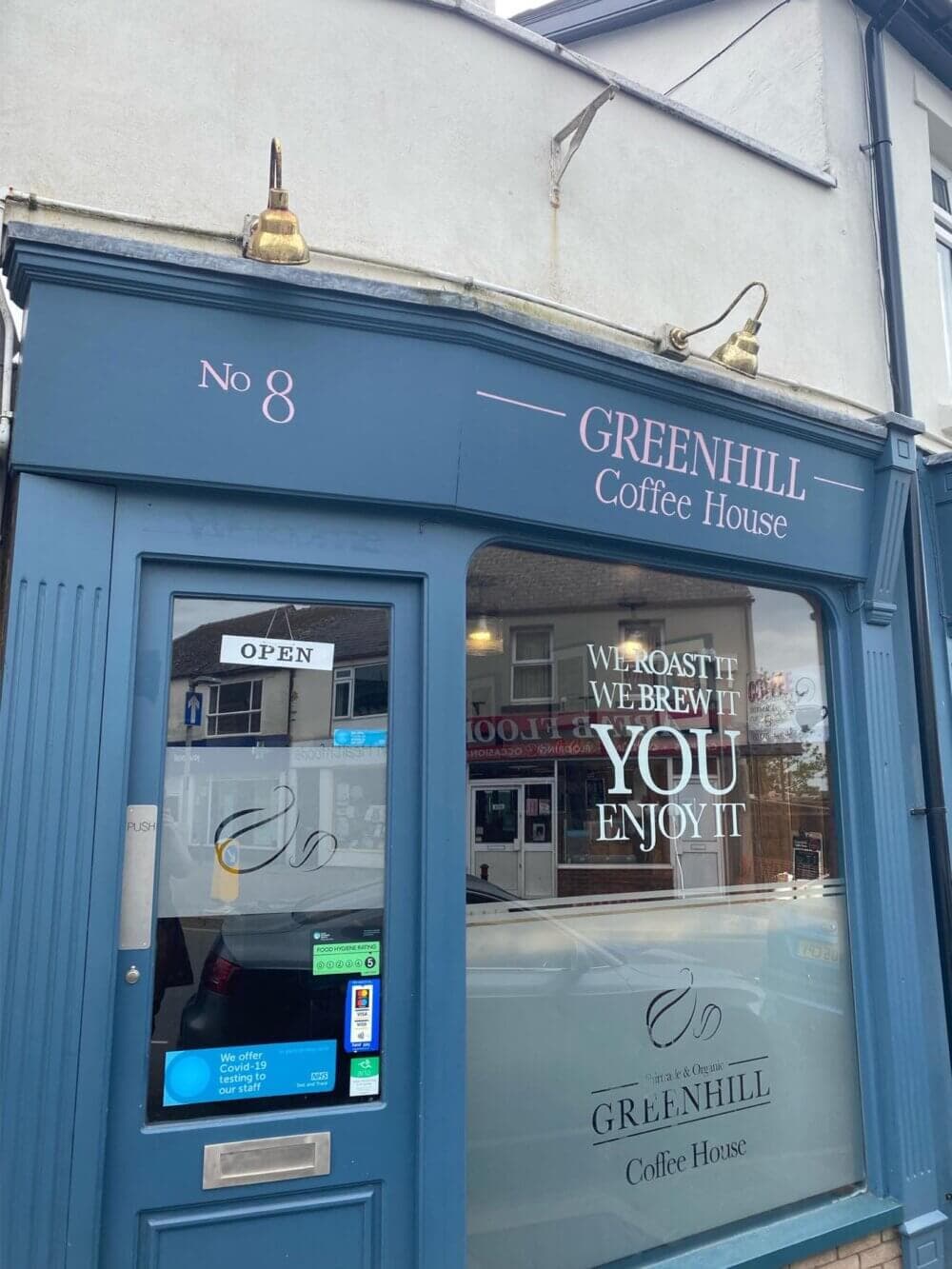 A blue storefront of Greenhill Coffee House with a "We roast it, we brew it, you enjoy it" sign on the window. - Home Instead