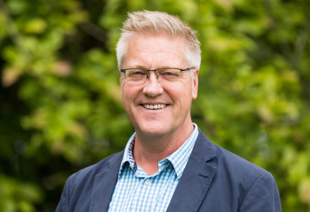 Smiling person with glasses, wearing a blue checkered shirt and blue blazer, against a blurred green leafy background. - Home Instead