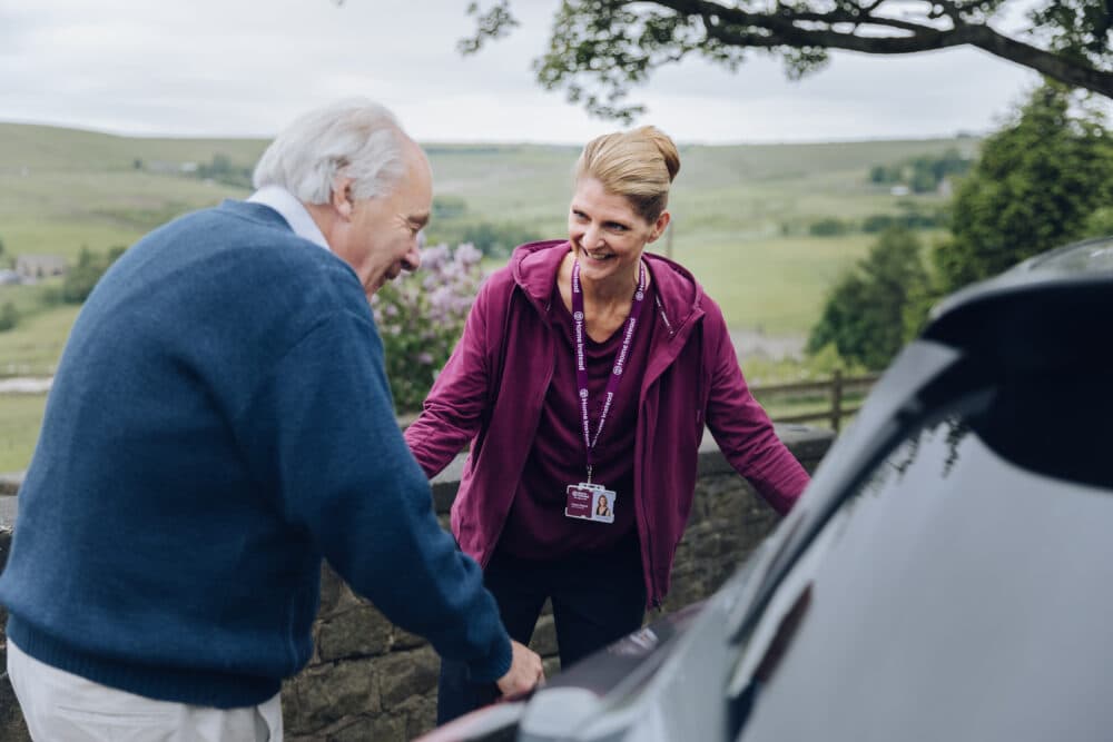 A caregiver assists an elderly man into a car outdoors, both smiling, with a scenic countryside background. - Home Instead