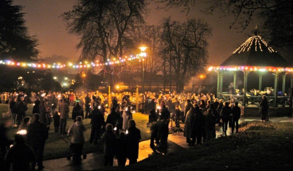 A large crowd gathers around a lit gazebo under string lights at night, holding candles in a park setting. - Home Instead