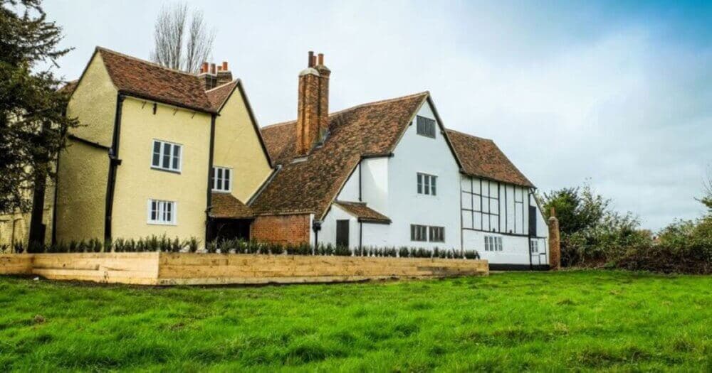 Historic two-story house with two chimneys, white and beige walls, and a sloped roof surrounded by green grass. - Home Instead