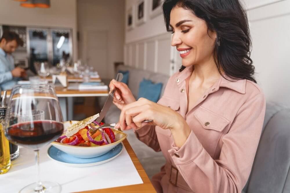 Woman in a pink blouse dining in a restaurant, smiling while holding a fork and knife over a dish, with wine glass nearby. - Home Instead