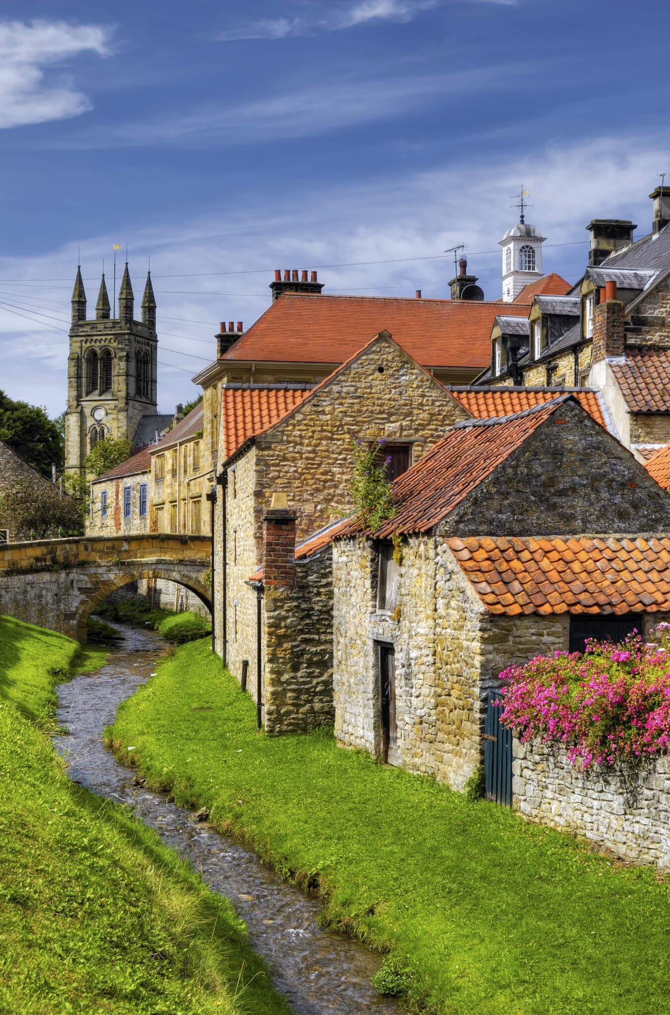 A quaint village with stone buildings, a small stream, and a church with tall spires in the background under a blue sky. - Home Instead