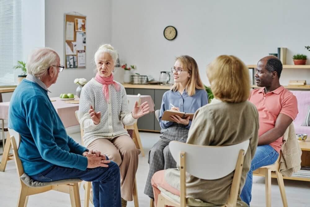 A group of people sits in a circle in a brightly lit room, engaged in a discussion. - Home Instead