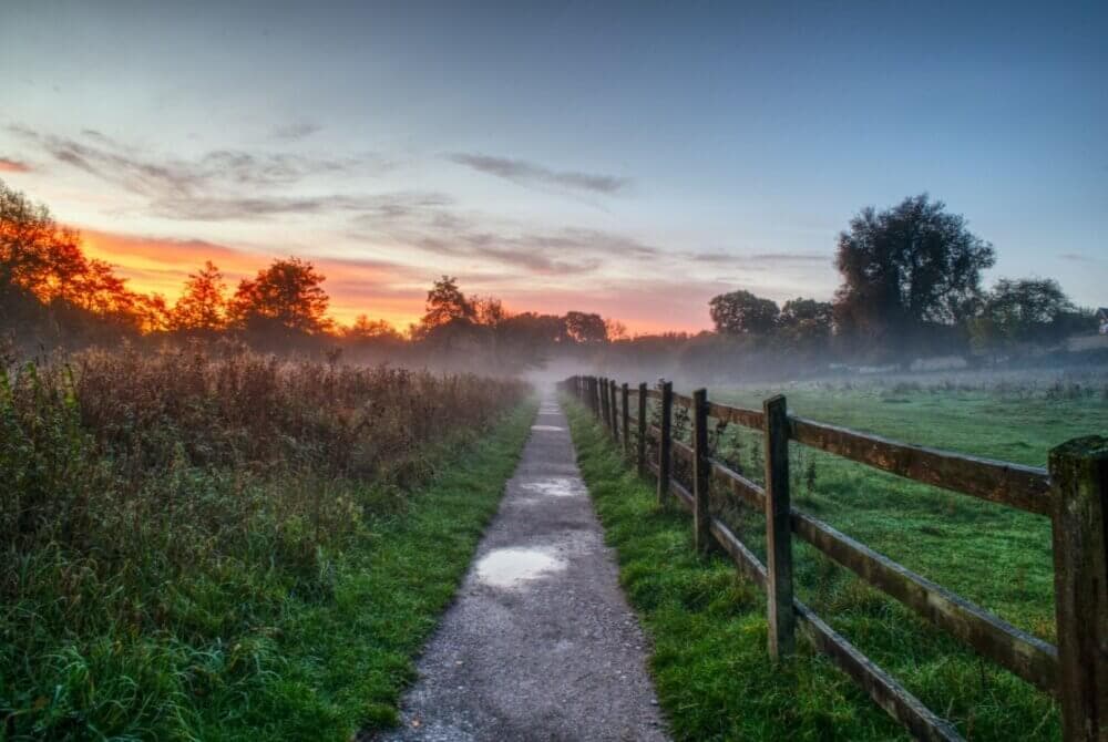 A rural pathway flanked by fields and a wooden fence, with a misty sunrise and colorful sky in the background. - Home Instead