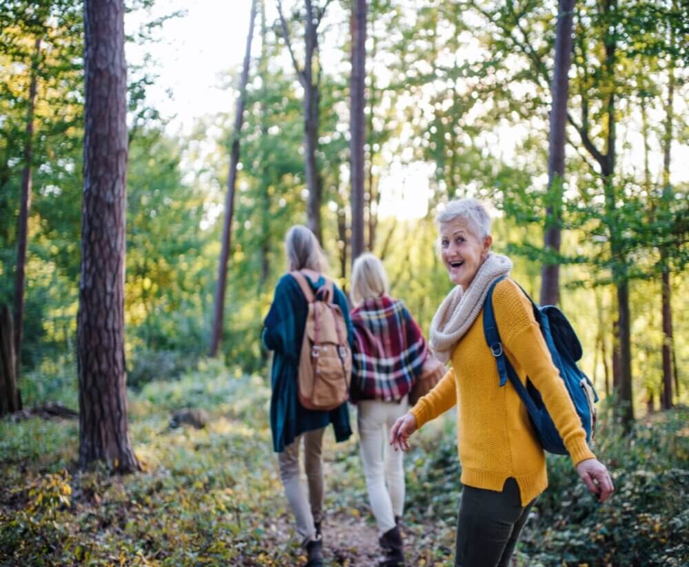 Three women hiking through a forest, one smiling and looking back. They are wearing backpacks and casual outdoor clothing. - Home Instead