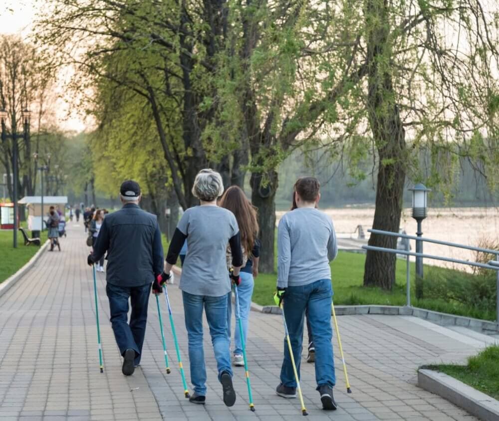 Four people walking with poles on a tree-lined path beside a river in the evening. - Home Instead