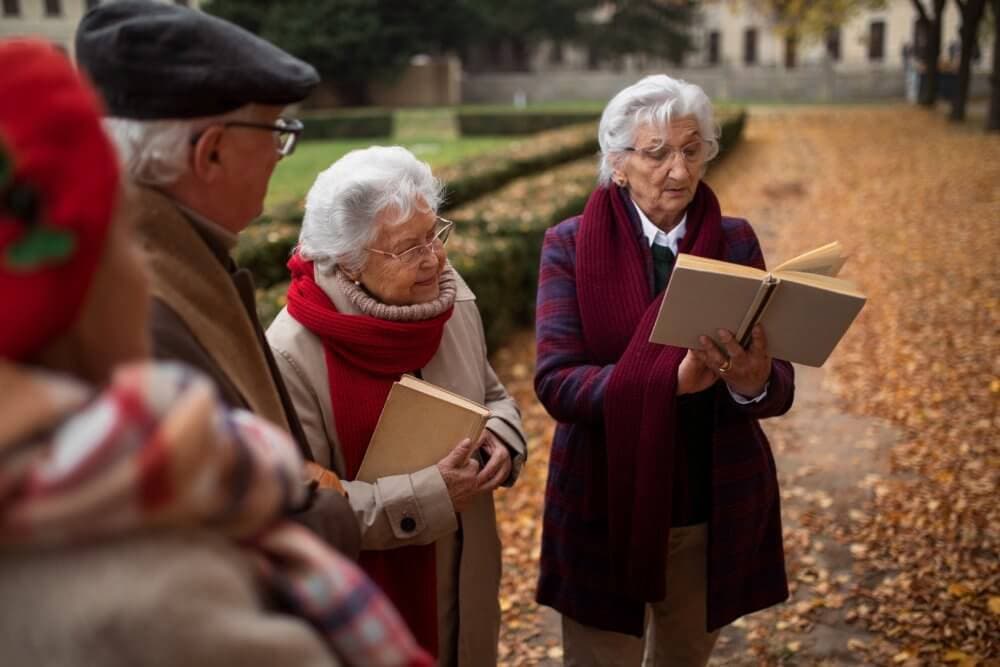 A group of elderly people standing outdoors; one person is reading a book, surrounded by autumn leaves. - Home Instead