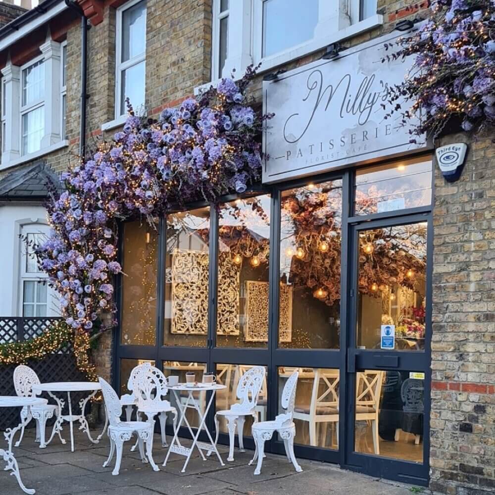 A quaint patisserie with a floral façade, white outdoor furniture, and large windows revealing a charming interior. - Home Instead