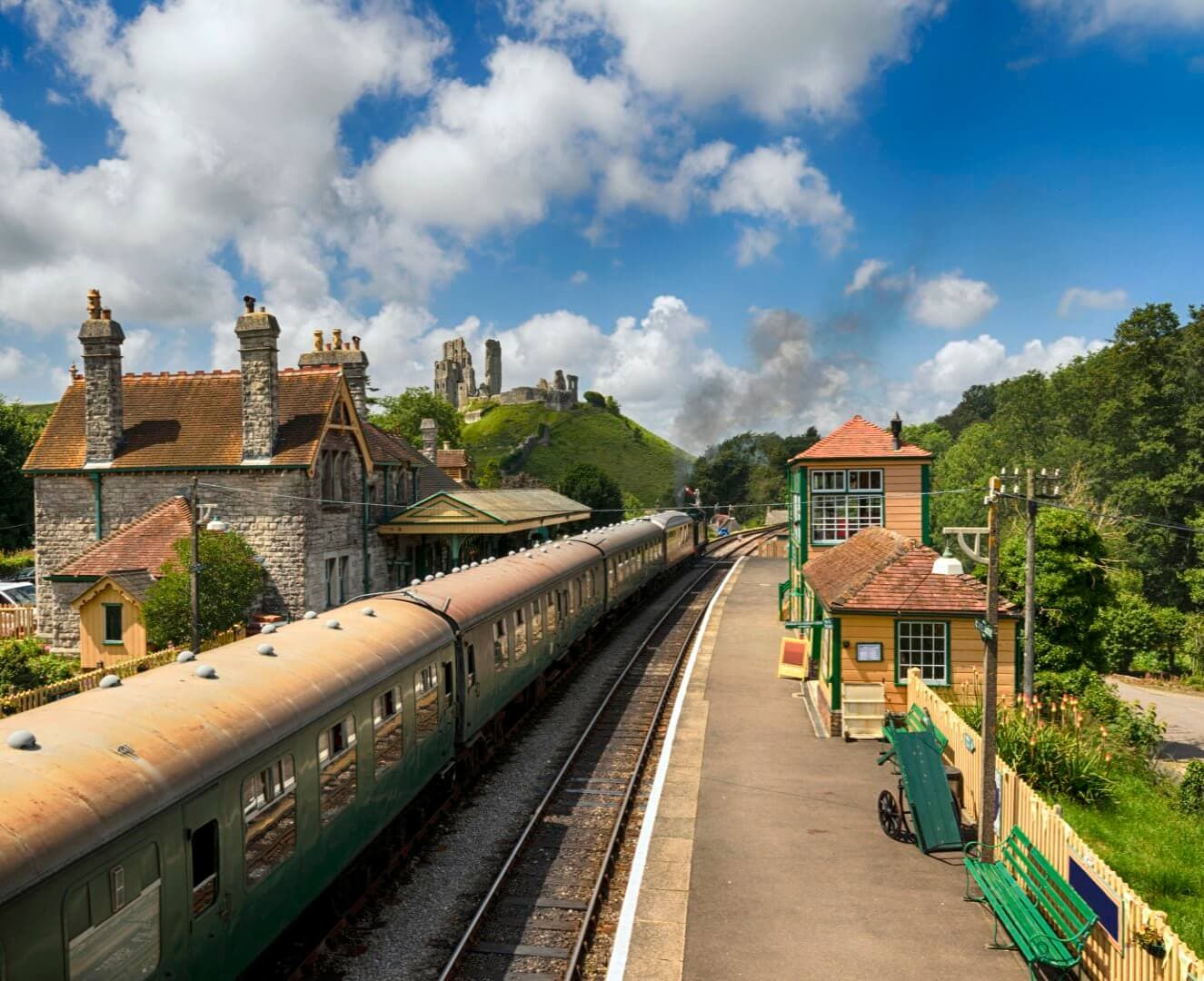 A vintage train arrives at a quaint countryside station with historic castle ruins visible in the green hills beyond. - Home Instead Poole