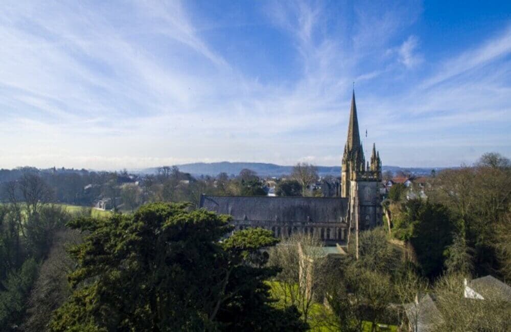 Aerial view of a historic church with tall spires surrounded by lush greenery and distant hills under a blue sky. - Home Instead