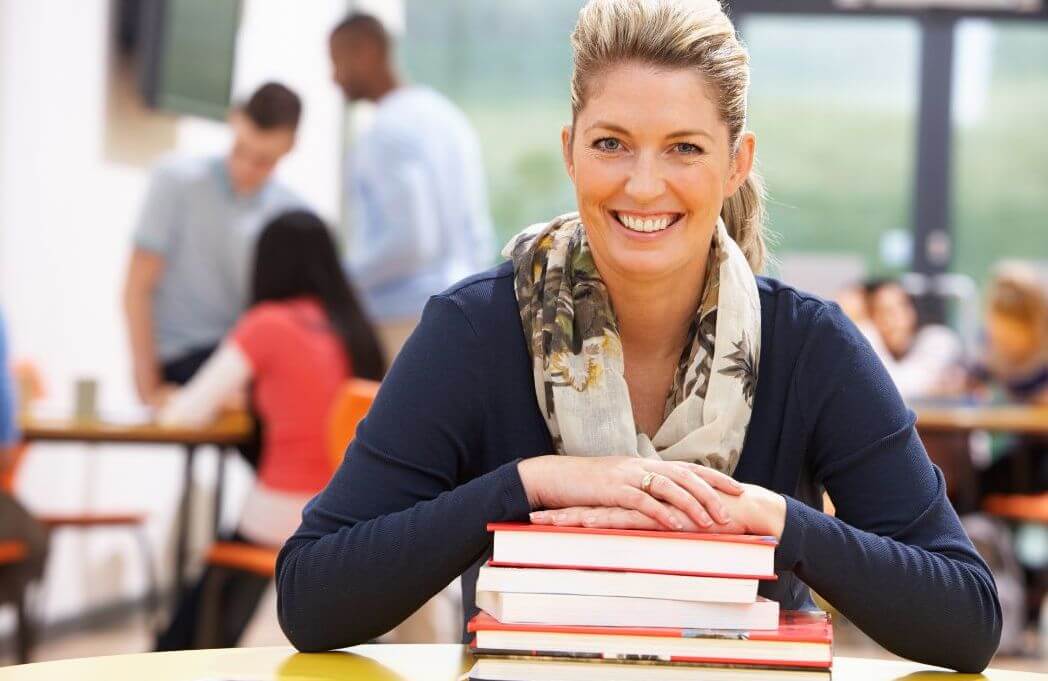 Smiling woman in a classroom rests her arms on a stack of books, with students interacting in the background. - Home Instead